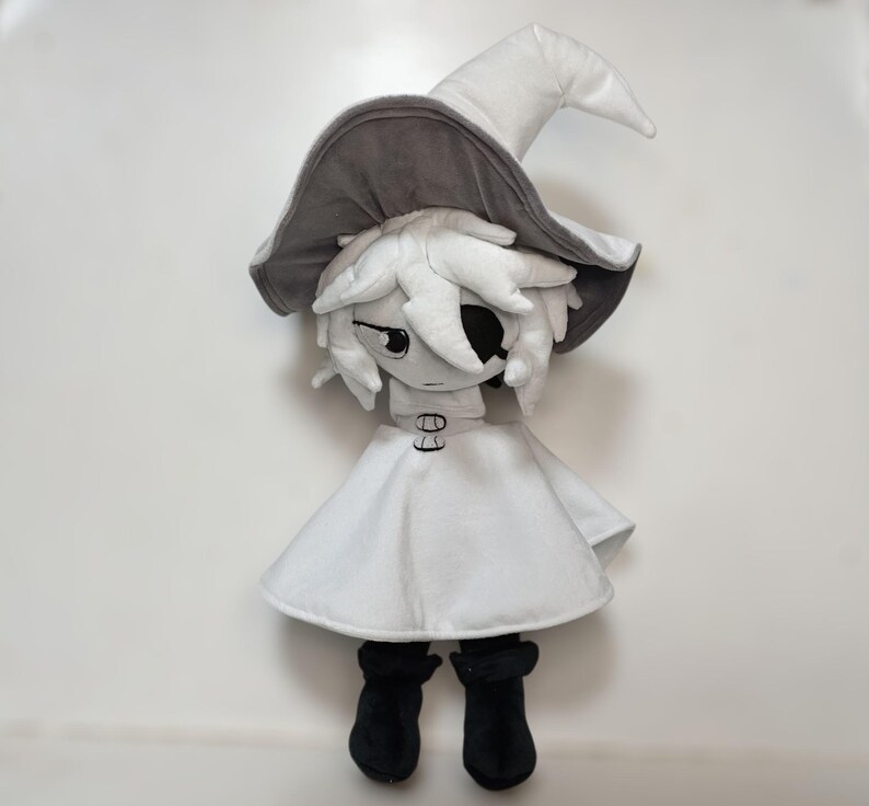 K&ouml;nnte beinhalten: Pl&uuml;schpuppe einer Figur mit wei&szlig;em Kleid, schwarzen Stiefeln und einem grau-wei&szlig;en Hexenhut. Die Puppe hat wei&szlig;es Haar und eine schwarze Augenklappe. Die Puppe steht aufrecht vor einem wei&szlig;en Hintergrund.
