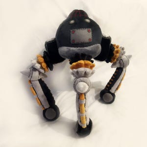 May include: A handmade plush toy robot in shades of grey, black, and yellow. The robot has a rounded head with a red and black top, a grey body, and three articulated legs with yellow and black accents. The toy is made of soft fabric.