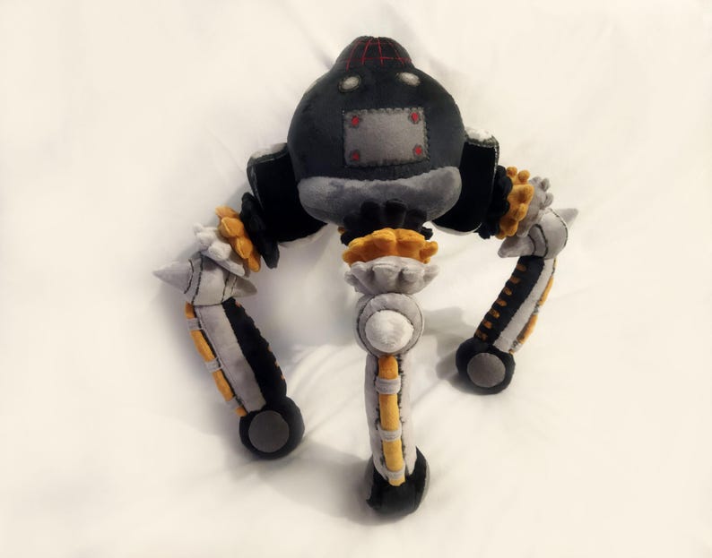 May include: A handmade plush toy robot in shades of grey, black, and yellow. The robot has a rounded head with a red and black top, a grey body, and three articulated legs with yellow and black accents. The toy is made of soft fabric.