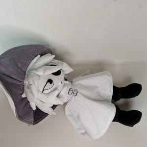 K&ouml;nnte beinhalten: Pl&uuml;schpuppe mit wei&szlig;em Gesicht, schwarzen Augen und einem aufgestickten L&auml;cheln. Die Puppe tr&auml;gt einen wei&szlig;en Umhang, schwarze Stiefel und eine graue Kapuze. Die Puppe liegt auf einer wei&szlig;en Oberfl&auml;che.