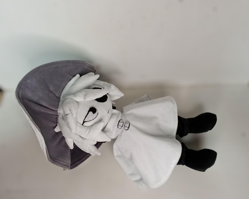 K&ouml;nnte beinhalten: Pl&uuml;schpuppe mit wei&szlig;em Gesicht, schwarzen Augen und einem aufgestickten L&auml;cheln. Die Puppe tr&auml;gt einen wei&szlig;en Umhang, schwarze Stiefel und eine graue Kapuze. Die Puppe liegt auf einer wei&szlig;en Oberfl&auml;che.