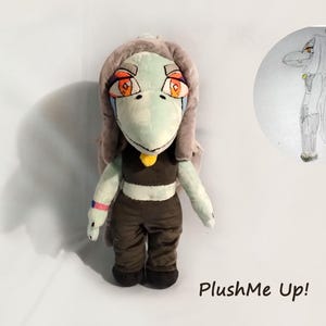 May include: A soft toy doll with a pale green face, large orange eyes, and grey hair. The doll is dressed in a brown crop top and trousers. A drawing of the doll is in a circle on the right. The text "PlushMe Up!" is at the bottom.