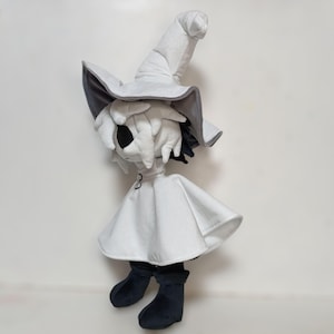 K&ouml;nnte beinhalten: Pl&uuml;schpuppe einer Figur mit einem wei&szlig;en Hexenhut mit grauem Rand, einem wei&szlig;en Umhang und schwarzen Stiefeln. Die Puppe hat ein wei&szlig;es Gesicht mit schwarzen Augen und schwarzem Haar, das unter dem Hut hervorblitzt.