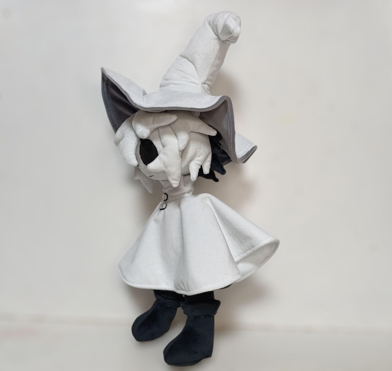 K&ouml;nnte beinhalten: Pl&uuml;schpuppe einer Figur mit einem wei&szlig;en Hexenhut mit grauem Rand, einem wei&szlig;en Umhang und schwarzen Stiefeln. Die Puppe hat ein wei&szlig;es Gesicht mit schwarzen Augen und schwarzem Haar, das unter dem Hut hervorblitzt.