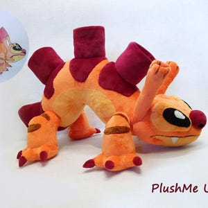 May include: A plush toy resembling a creature with orange fur, dark red spots, and dark red dorsal plates. The character has large black eyes, a small nose, and a single visible tooth. A drawing of the character is in the top left corner.