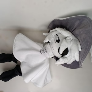 K&ouml;nnte beinhalten: Pl&uuml;schpuppe in Form einer Zeichentrickfigur mit wei&szlig;em Kopf und H&auml;nden, wei&szlig;em Outfit und schwarzen Schuhen. Die Puppe hat einen grauen Hut und ein gro&szlig;es, ausdrucksstarkes Gesicht mit schwarzen Augen und einem breiten L&auml;cheln.