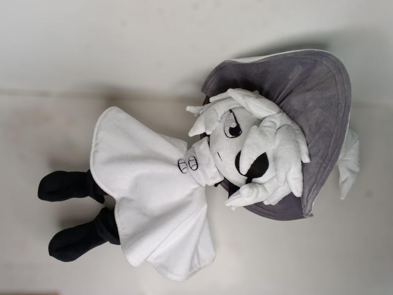 K&ouml;nnte beinhalten: Pl&uuml;schpuppe in Form einer Zeichentrickfigur mit wei&szlig;em Kopf und H&auml;nden, wei&szlig;em Outfit und schwarzen Schuhen. Die Puppe hat einen grauen Hut und ein gro&szlig;es, ausdrucksstarkes Gesicht mit schwarzen Augen und einem breiten L&auml;cheln.
