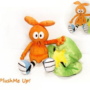May include: An orange plush toy with big eyes, long ears, and outstretched arms, next to a green hat with a yellow star. The toy has blue and white striped shoes. The text "PlushMe Up!" is in the bottom left corner.