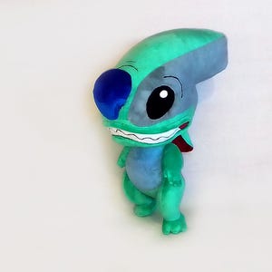 Peut inclure: Une peluche repr&eacute;sentant un personnage de dessin anim&eacute;, avec une t&ecirc;te turquoise et grise, de grands yeux noirs et une oreille bleue. Le personnage a un large sourire avec des dents blanches et une langue rouge, et un corps bleu clair. La peluche est sur une surface blanche.