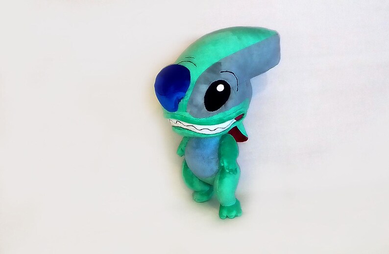 Peut inclure: Une peluche repr&eacute;sentant un personnage de dessin anim&eacute;, avec une t&ecirc;te turquoise et grise, de grands yeux noirs et une oreille bleue. Le personnage a un large sourire avec des dents blanches et une langue rouge, et un corps bleu clair. La peluche est sur une surface blanche.