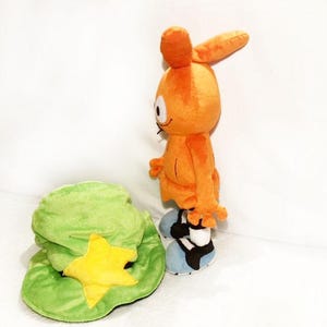 May include: An orange plush toy character with large ears, standing next to a green hat with a yellow star. The character has blue and black shoes. The plush toy is a cartoon character.