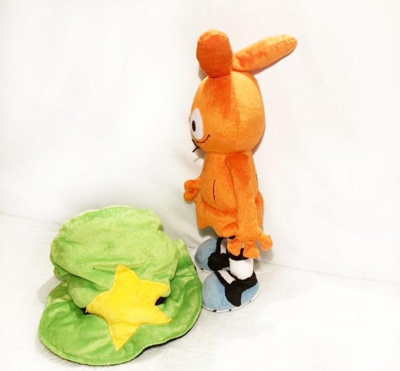 May include: An orange plush toy character with large ears, standing next to a green hat with a yellow star. The character has blue and black shoes. The plush toy is a cartoon character.
