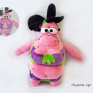 May include: Pink plush toy resembling a cartoon character, with a grumpy expression and large eyes. It wears a purple and green outfit. A drawing of the character is in the upper left corner. The text "PlushMe Up!" is in the lower right corner.