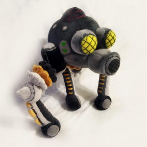 May include: A soft toy robot in grey, black, and yellow. The robot features a rounded head with a red stripe, two large yellow-grid eyes, and multiple articulated limbs. The toy is made from soft, textured materials.