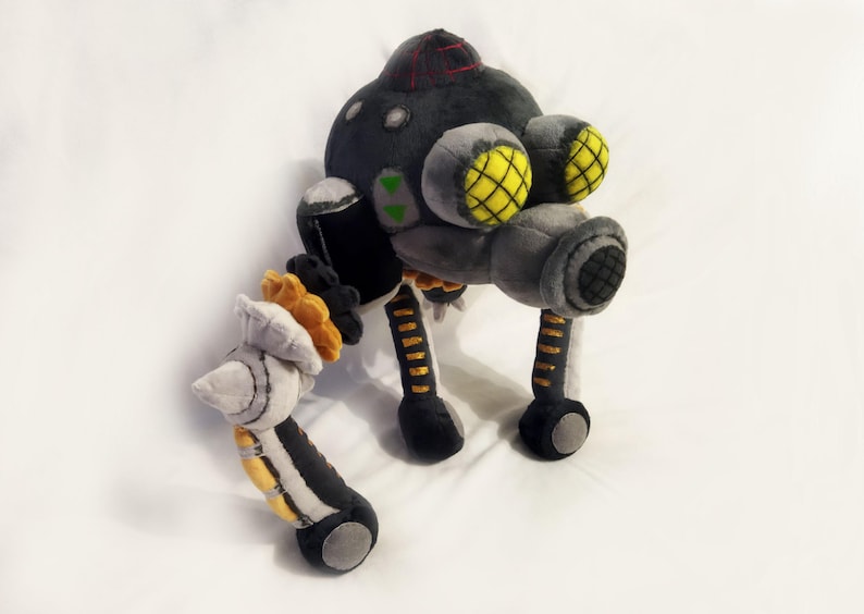 May include: A soft toy robot in grey, black, and yellow. The robot features a rounded head with a red stripe, two large yellow-grid eyes, and multiple articulated limbs. The toy is made from soft, textured materials.