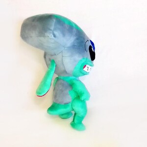 Peut inclure: Peluche repr&eacute;sentant un extraterrestre gris et turquoise. L'alien a de grandes oreilles, une grosse t&ecirc;te et un large sourire avec des dents visibles. Le jouet est doux et c&acirc;lin, pour enfants ou collectionneurs.