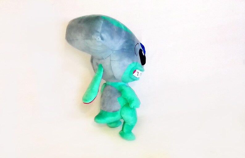 Peut inclure: Peluche repr&eacute;sentant un extraterrestre gris et turquoise. L'alien a de grandes oreilles, une grosse t&ecirc;te et un large sourire avec des dents visibles. Le jouet est doux et c&acirc;lin, pour enfants ou collectionneurs.
