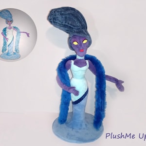 Peut inclure: Une poupée en peluche représentant un personnage à la peau violette avec de hauts cheveux bleu foncé, des yeux jaunes et des lèvres rouges. La poupée porte une robe bleu clair avec une bordure bleu foncé et un long boa bleu duveteux.