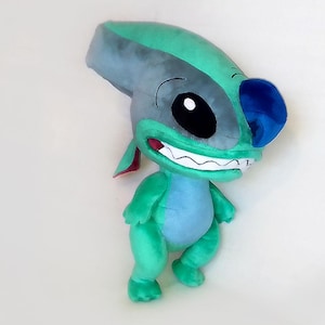 Peut inclure: Peluche repr&eacute;sentant un personnage de dessin anim&eacute; avec un corps turquoise, une t&ecirc;te grise et des d&eacute;tails bleus. Le personnage a de grands yeux noirs, un large sourire et des oreilles pointues. Jouet doux.