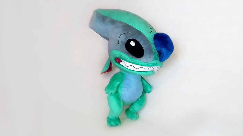 Peut inclure: Peluche repr&eacute;sentant un personnage de dessin anim&eacute; avec un corps turquoise, une t&ecirc;te grise et des d&eacute;tails bleus. Le personnage a de grands yeux noirs, un large sourire et des oreilles pointues. Jouet doux.