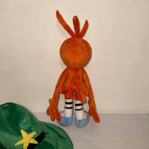 May include: An orange plush toy with long arms and legs, white socks with black stripes, and blue shoes. A green hat with a yellow star and black band is in the foreground. The toy has a round head with three orange antennae.