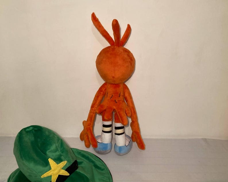 May include: An orange plush toy with long arms and legs, white socks with black stripes, and blue shoes. A green hat with a yellow star and black band is in the foreground. The toy has a round head with three orange antennae.