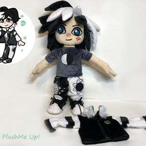 May include: A black and white plush toy with black hair, a grey shirt with a crescent moon design, and black and white pants with a sun and moon design. The toy is wearing a black jacket with a green alien design and white and black striped leg warmers. The toy is standing on a white surface. PlushMe Up! is written on the surface.