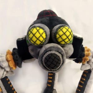May include: A plush toy robot with a black head and body, grey face and arms, and yellow eyes with black grid patterns. The robot has a grey respirator and orange and white accents.