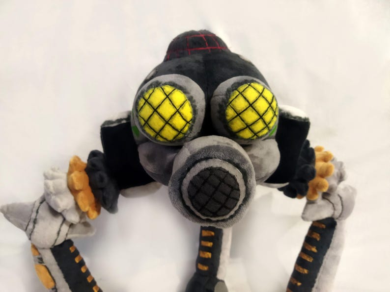May include: A plush toy robot with a black head and body, grey face and arms, and yellow eyes with black grid patterns. The robot has a grey respirator and orange and white accents.