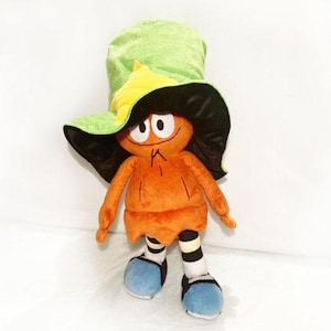 May include: A soft toy of a cartoon character with an orange body, black hair, and a green and yellow hat. The toy has blue shoes and striped socks. The character has large eyes and a small mouth.