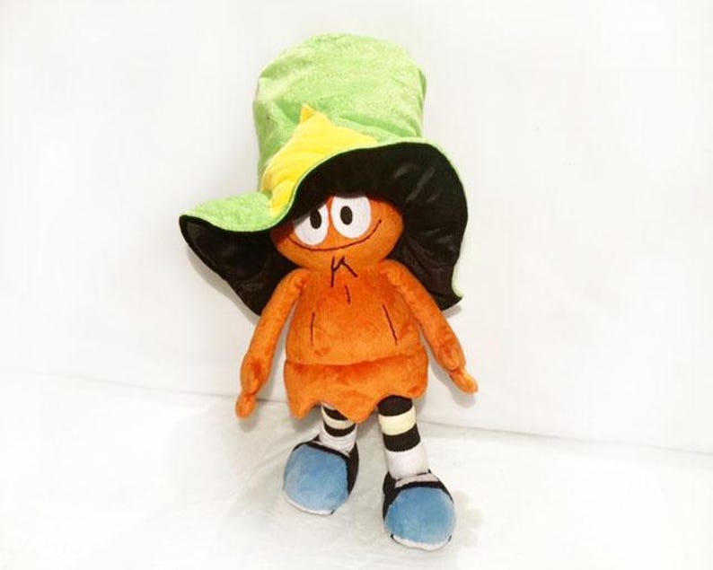 May include: A soft toy of a cartoon character with an orange body, black hair, and a green and yellow hat. The toy has blue shoes and striped socks. The character has large eyes and a small mouth.