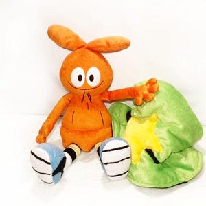 May include: A soft orange toy with large eyes, rabbit-like ears, and a small smile. It has orange arms and legs with blue and white details. A green hat with a yellow star is next to the toy.