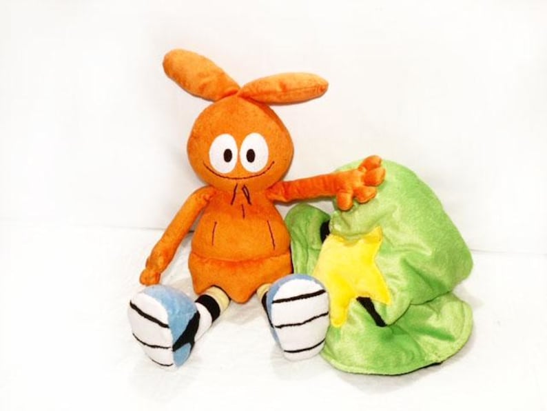 May include: A soft orange toy with large eyes, rabbit-like ears, and a small smile. It has orange arms and legs with blue and white details. A green hat with a yellow star is next to the toy.
