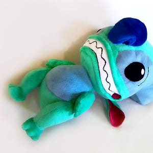 Peut inclure: Une peluche en forme de personnage de dessin anim&eacute;, avec une t&ecirc;te bleue, de grands yeux noirs, un visage turquoise et une langue rouge. Le jouet a des bras et des jambes verts et un corps bleu. Le jouet est pos&eacute; sur une surface blanche.