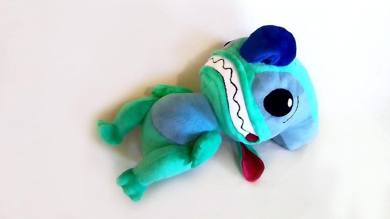 Peut inclure: Une peluche en forme de personnage de dessin anim&eacute;, avec une t&ecirc;te bleue, de grands yeux noirs, un visage turquoise et une langue rouge. Le jouet a des bras et des jambes verts et un corps bleu. Le jouet est pos&eacute; sur une surface blanche.