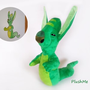 May include: A green plush toy creature with large ears and a long tail, alongside a watercolour illustration of the same character. The toy has a light green belly. The text "PlushMe Up!" is visible in the image.