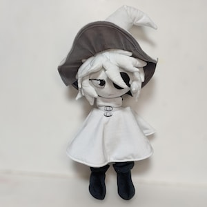 K&ouml;nnte beinhalten: Pl&uuml;schpuppe einer Figur, die ein wei&szlig;es Kleid, schwarze Stiefel und einen grau-wei&szlig;en Hexenhut tr&auml;gt. Die Puppe hat ein stilisiertes Gesicht mit schwarzen Augen und einem neutralen Ausdruck. Die Puppe ist aus weichem Stoff.