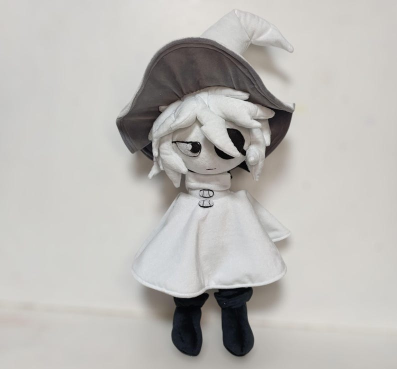 K&ouml;nnte beinhalten: Pl&uuml;schpuppe einer Figur, die ein wei&szlig;es Kleid, schwarze Stiefel und einen grau-wei&szlig;en Hexenhut tr&auml;gt. Die Puppe hat ein stilisiertes Gesicht mit schwarzen Augen und einem neutralen Ausdruck. Die Puppe ist aus weichem Stoff.