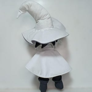 K&ouml;nnte beinhalten: Pl&uuml;schpuppe eines wei&szlig;en Magiers mit einem spitzen Hut und Umhang, kombiniert mit schwarzen Stiefeln. Das Gesicht der Puppe ist von einer schwarzen Maske verdeckt. Das Design ist minimalistisch und betont den klassischen Magier-Look.