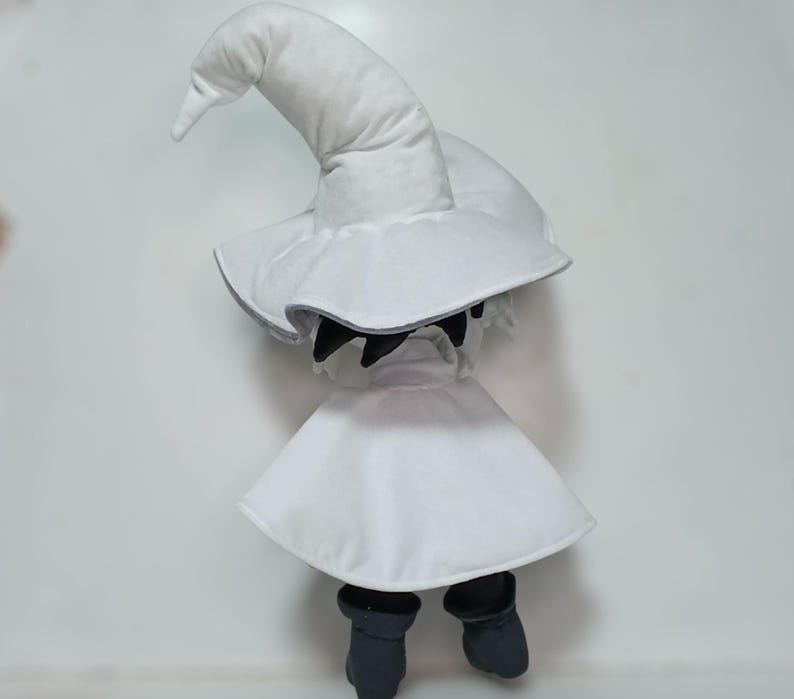 K&ouml;nnte beinhalten: Pl&uuml;schpuppe eines wei&szlig;en Magiers mit einem spitzen Hut und Umhang, kombiniert mit schwarzen Stiefeln. Das Gesicht der Puppe ist von einer schwarzen Maske verdeckt. Das Design ist minimalistisch und betont den klassischen Magier-Look.
