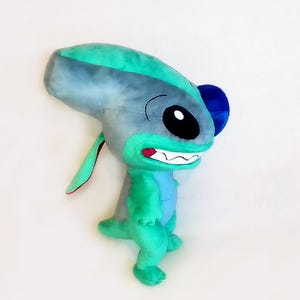 Peut inclure: Peluche repr&eacute;sentant un personnage extraterrestre bleu et vert, avec de grands yeux noirs et un large sourire. Le jouet a de grandes oreilles et une petite queue, sur fond blanc.