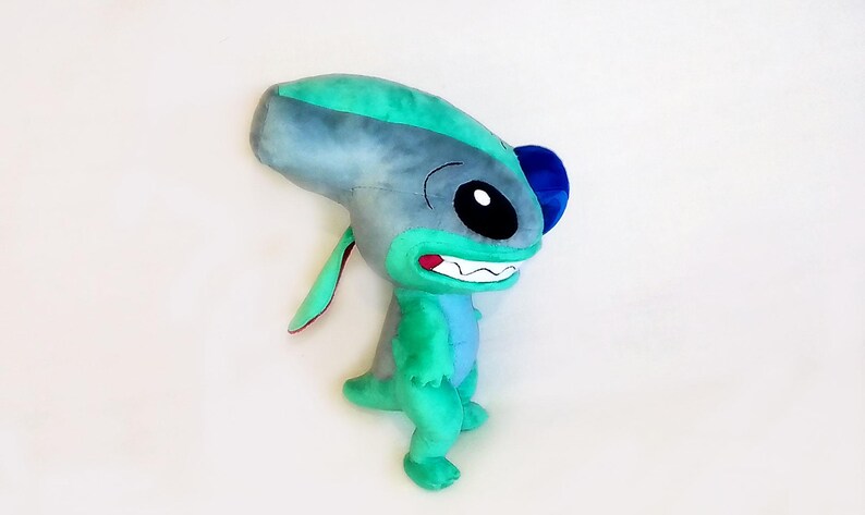 Peut inclure: Peluche repr&eacute;sentant un personnage extraterrestre bleu et vert, avec de grands yeux noirs et un large sourire. Le jouet a de grandes oreilles et une petite queue, sur fond blanc.