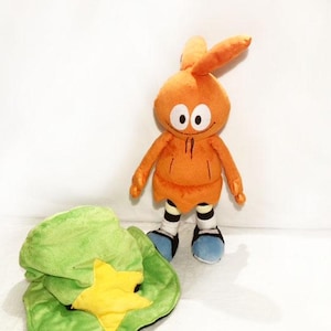 May include: An orange plush toy with large white eyes, a smiling face, and long ears. It wears a hooded outfit and blue shoes. A green plush hat with a yellow star is in the foreground.