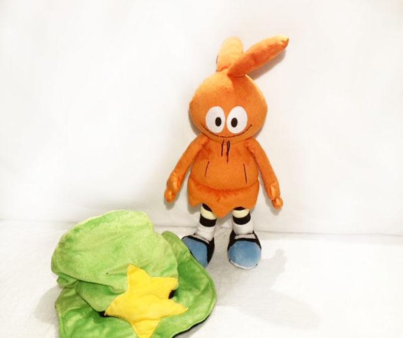 May include: An orange plush toy with large white eyes, a smiling face, and long ears. It wears a hooded outfit and blue shoes. A green plush hat with a yellow star is in the foreground.