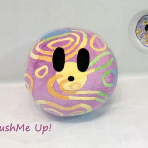 May include: A plush, spherical toy with a face, featuring a light purple base color and colorful swirls in yellow, green, and blue. The toy has large black eyes and a small black mouth. A smaller, hand-drawn version is also visible.