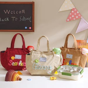 Puede incluir: Tres bolsos de mano de pana en rojo, beige y marrón, cada uno con un nombre bordado en la parte delantera. Los bolsos tienen asas y detalles florales. También se ven una fiambrera y una botella de agua.