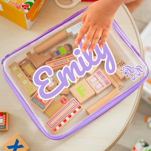 May include: A clear plastic case with a purple zipper and trim, filled with wooden toy blocks. The blocks include buildings, people, and signs. A large, purple sticker with the name "Emily" is on the case, along with a unicorn sticker.