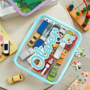 May include: A clear, rectangular storage case with a light blue zipper, filled with various toy cars. The name "Oliver" is written in a cursive font across the front. A yellow vintage car is visible outside the case.