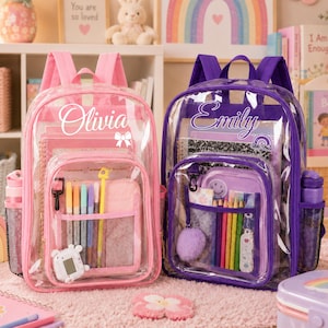 May include: Two clear backpacks, one pink and one purple, featuring personalised names Olivia and Emily in white script. Each bag has a front pocket with mesh organisers containing coloured pencils, pens, and small accessories like a pom-pom.