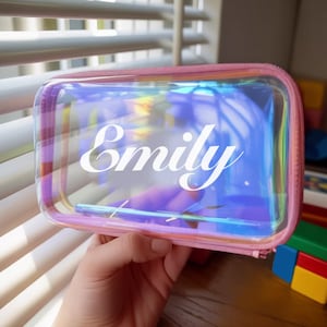 Może przedstawiać: Przezroczysta, opalizująca kosmetyczka z różowym zamkiem błyskawicznym i wykończeniem. Imię "Emily" jest wydrukowane białym skryptem na przodzie. Torba jest prostokątna i wydaje się być wykonana z przezroczystego materiału.